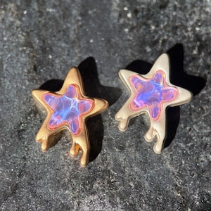 May include: Two star-shaped pins featuring a matte gold finish with a dripping, melted effect. Each star has a central iridescent, translucent inlay that shifts between shades of pink and blue, creating a glowing, liquid-like appearance.