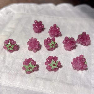 May include: A collection of ten small, three-dimensional raspberry-shaped beads. Each bead is crafted from translucent pink glass with a cluster of rounded segments and features a small, light green painted leaf detail at the base of the fruit.