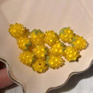 May include: A collection of small, translucent yellow glass beads shaped like raspberries. Each bead features a textured, bumpy surface and is topped with a small, green-striped cap detail, mimicking the appearance of individual fruit segments.