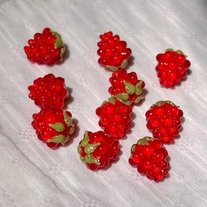 May include: A collection of nine small, translucent red raspberry-shaped beads. Each bead features a textured, bumpy surface resembling individual drupelets and includes small, light green painted leaf accents at the base of the fruit.