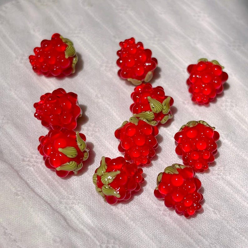 May include: A collection of nine small, translucent red raspberry-shaped beads. Each bead features a textured, bumpy surface resembling individual drupelets and includes small, light green painted leaf accents at the base of the fruit.