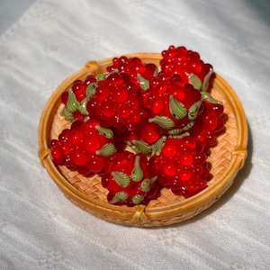 May include: A small, round, woven bamboo basket filled with several bright red glass raspberries. Each raspberry features a translucent, bumpy texture and small, light green glass leaves attached to the base of the fruit.