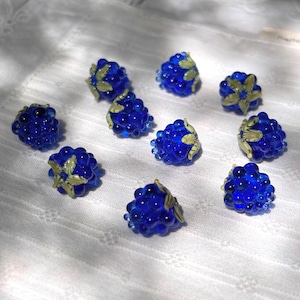 May include: A collection of nine individual glass beads shaped like clusters of berries. Each bead is a deep, translucent cobalt blue with small, raised spherical textures and a pale green leaf detail attached to the top of each berry cluster.