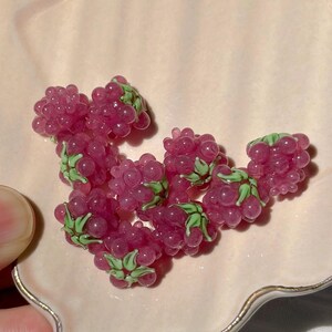 May include: A collection of small, translucent raspberry-shaped beads made of glass. Each bead features a cluster of pink, rounded segments with a small, light green painted leaf detail at the base, creating a realistic fruit appearance.