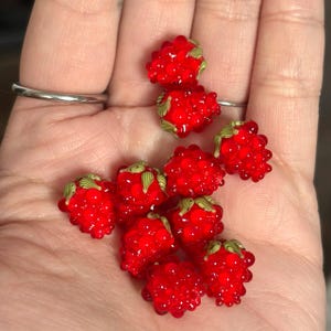 May include: A collection of small, bright red glass beads shaped like raspberries. Each bead features a textured, bumpy surface and is topped with a small, detailed green glass leaf element. The beads are translucent and have a glossy finish.