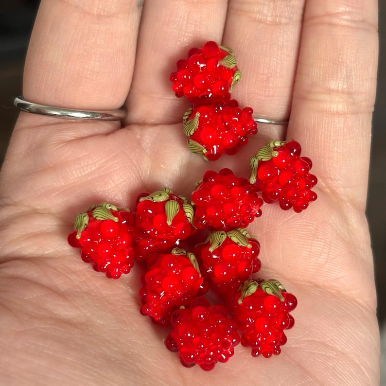 May include: A collection of small, bright red glass beads shaped like raspberries. Each bead features a textured, bumpy surface and is topped with a small, detailed green glass leaf element. The beads are translucent and have a glossy finish.