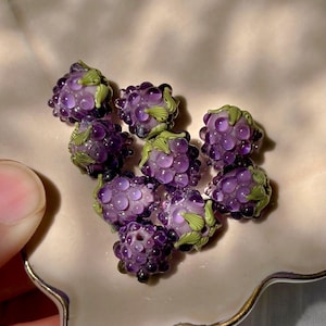 May include: A collection of small, handcrafted glass beads shaped like clusters of purple grapes. Each bead features translucent purple spheres arranged in a bunch with small, detailed green glass leaves attached to the top of each cluster.