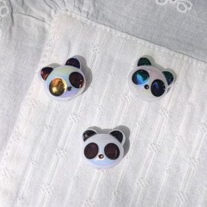 May include: Three small, white, panda-shaped cabochons with iridescent, reflective surfaces. Each panda features dark, circular eye patches and a small, centered nose detail. The finish on the panda faces shifts between blue, purple, and gold tones.