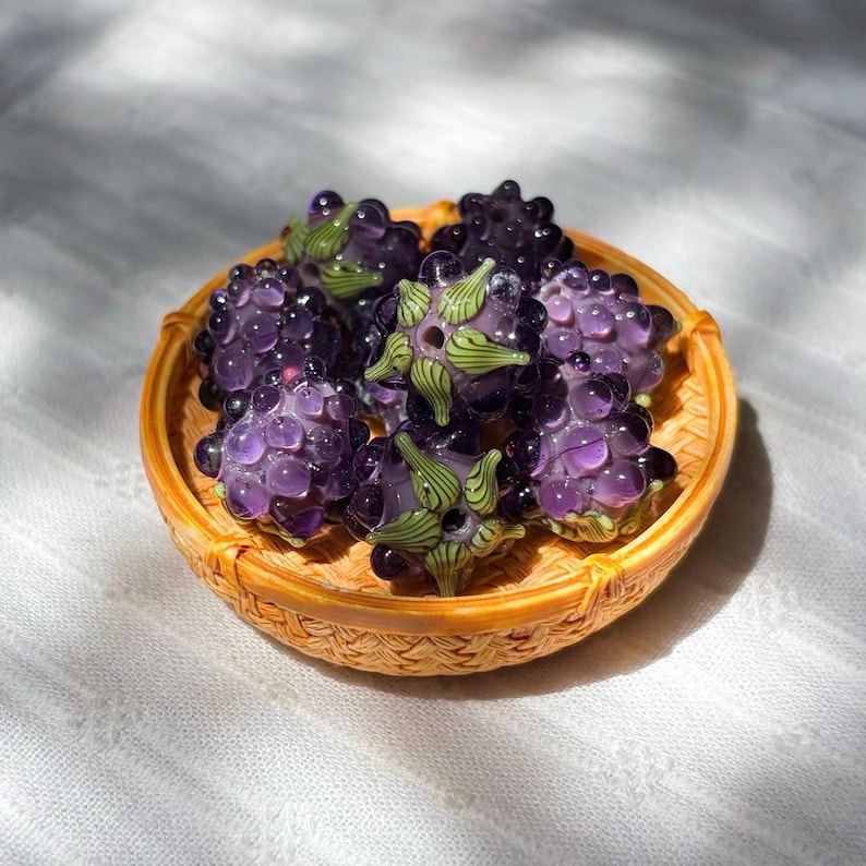 May include: A collection of translucent purple glass beads shaped like blackberries, each featuring a small green glass stem detail. The beads are clustered together inside a small, round, woven light brown basket with a textured rim.