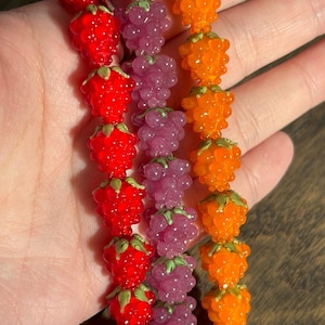 May include: Three strands of glass beads shaped like raspberries, arranged in vertical rows. The left strand is bright red, the middle strand is translucent purple, and the right strand is vibrant orange. Each bead features small green leaf details.