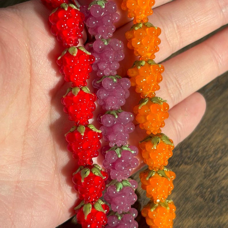 May include: Three strands of glass beads shaped like raspberries, arranged in vertical rows. The left strand is bright red, the middle strand is translucent purple, and the right strand is vibrant orange. Each bead features small green leaf details.