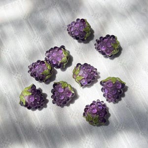 May include: A collection of eight individual glass beads shaped like clusters of purple grapes. Each bead features translucent, rounded purple segments and small, painted green leaf details at the top, creating a realistic fruit appearance.