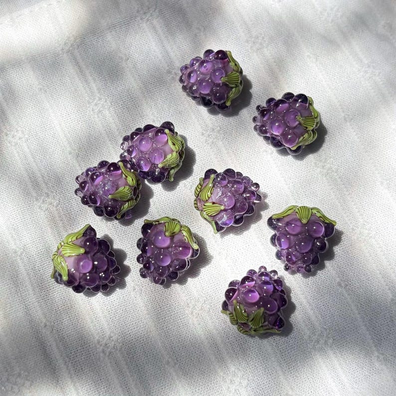 May include: A collection of eight individual glass beads shaped like clusters of purple grapes. Each bead features translucent, rounded purple segments and small, painted green leaf details at the top, creating a realistic fruit appearance.