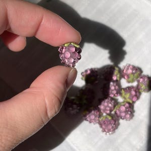 May include: A collection of small, translucent purple glass beads shaped like raspberries. Each bead features a cluster of rounded segments and a small green leaf detail at the top. The glass has a glossy finish that catches the light.