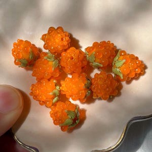 May include: A collection of ten small, translucent orange glass beads shaped like raspberries. Each bead features a textured, bumpy surface and is accented with small, light green glass leaves at the base, mimicking the appearance of fresh fruit.