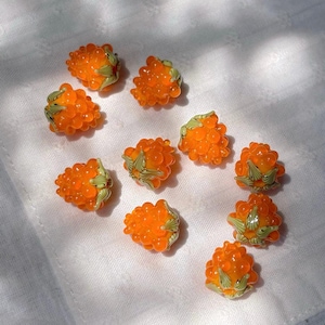 May include: A collection of ten small, translucent orange glass beads shaped like raspberries. Each bead features a textured, bumpy surface and is accented with small, light green glass leaves at the base, mimicking the appearance of fresh fruit.