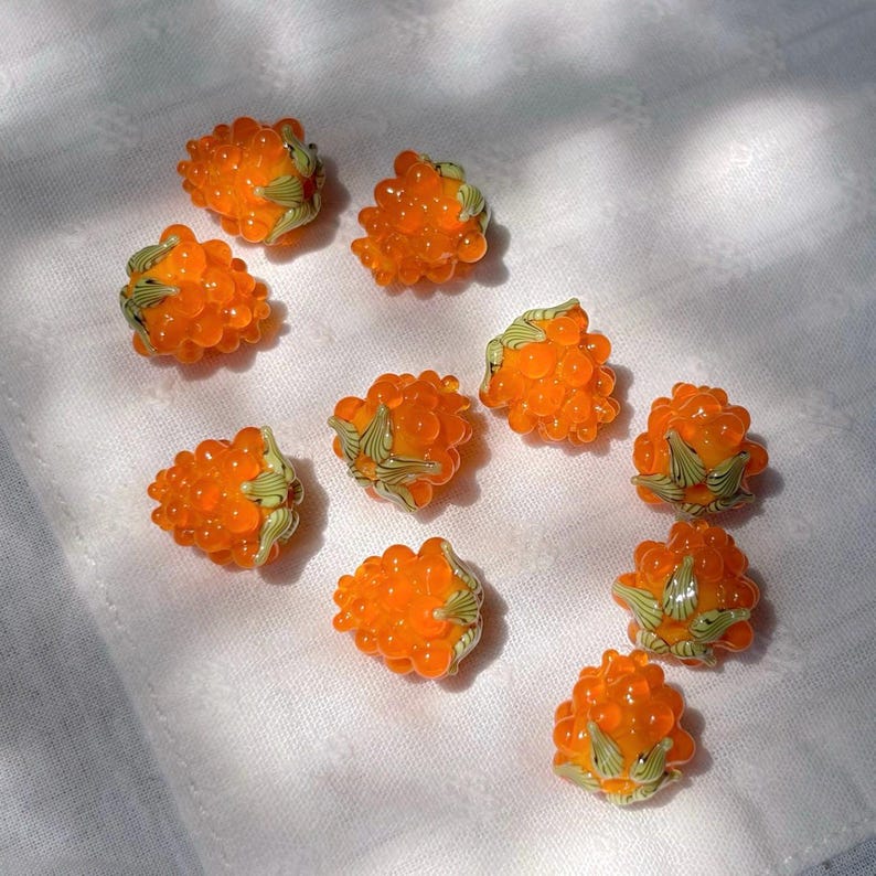 May include: A collection of ten small, translucent orange glass beads shaped like raspberries. Each bead features a textured, bumpy surface and is accented with small, light green glass leaves at the base, mimicking the appearance of fresh fruit.