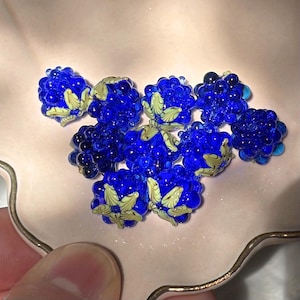 May include: A collection of nine cobalt blue glass beads shaped like blackberries, each featuring a textured surface and a small, pale yellow-green glass leaf detail attached to the top of the berry-shaped bead.
