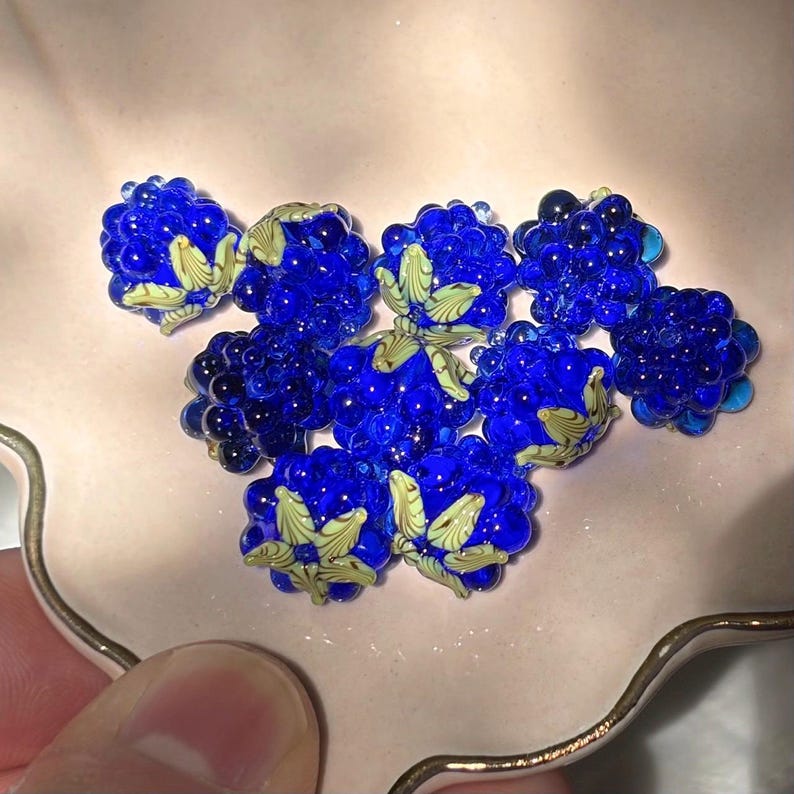 May include: A collection of nine cobalt blue glass beads shaped like blackberries, each featuring a textured surface and a small, pale yellow-green glass leaf detail attached to the top of the berry-shaped bead.