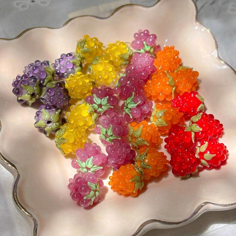 May include: A collection of small, translucent berry-shaped beads in vibrant colors including red, orange, yellow, pink, and purple. Each bead features a textured, bumpy surface resembling a raspberry or blackberry, topped with a small green cap.