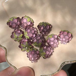 May include: A collection of small, translucent purple glass beads shaped like raspberries. Each bead features a cluster of rounded segments and a small, detailed green glass leaf element attached to the top, mimicking the appearance of fresh fruit.