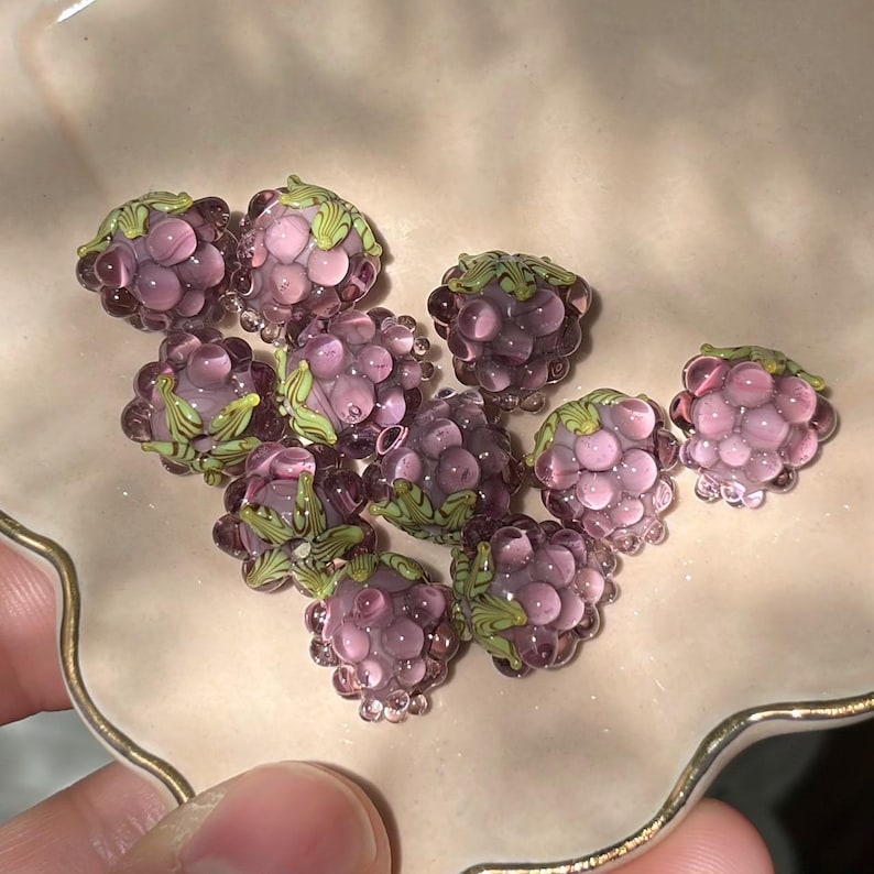 May include: A collection of small, translucent purple glass beads shaped like raspberries. Each bead features a cluster of rounded segments and a small, detailed green glass leaf element attached to the top, mimicking the appearance of fresh fruit.