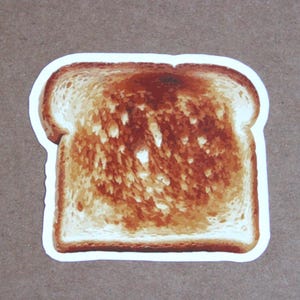 May include: A die-cut sticker shaped like a single slice of toasted bread. The toast features a gradient of golden-brown and dark brown toasted areas across the center, with lighter, un-toasted crust edges and a thin white border outline.