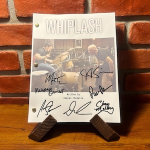 Peut inclure: Un scénario signé du film "Whiplash" présenté sur un présentoir en bois. Le script présente une photo d'un batteur et d'un homme, avec plusieurs signatures. L'arrière-plan est un mur de briques.