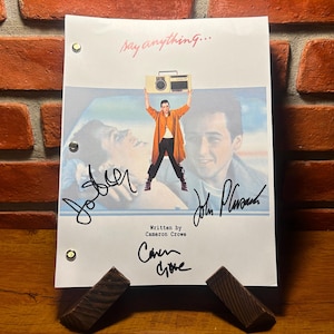 May include: A movie script for "Say Anything..." featuring a movie still of a man holding a boombox. The script is signed by the actors and the writer. The script is propped up on a wooden stand against a brick wall.