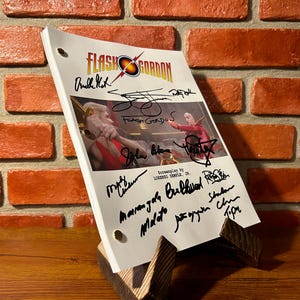 May include: A film script for "Flash Gordon" featuring the film's title and logo, along with multiple signatures. The script is displayed on a wooden stand, set against a backdrop of a brick wall.