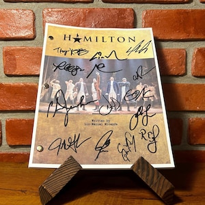 May include: A signed "Hamilton" playbill, featuring the show's title and cast signatures. The playbill is displayed on a wooden stand against a brick wall background. The text "Written by Lin-Manuel Miranda" is visible.