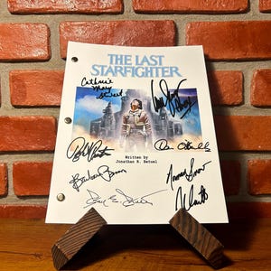 May include: A script for "The Last Starfighter" with multiple signatures, displayed on a wooden stand. The script's cover features a movie poster image of a man in a spacesuit. The background is a brick wall.