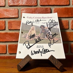 May include: A script from the movie "Vicky Cristina Barcelona" with multiple signatures, displayed on a wooden stand. The script is white with black text and images. The background is a brick wall.