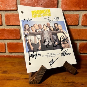 Puede incluir: Un guion firmado de Brooklyn Nine-Nine sobre un soporte de madera. El guion presenta el título del programa en amarillo y las firmas del elenco. El guion está apoyado en un soporte de madera contra una pared de ladrillos.