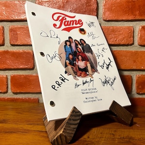 Op de afbeelding: Een script van de tv-show "Fame" op een houten standaard. Het script is wit met het logo van de show en een foto van de cast. Het is door meerdere mensen gesigneerd. De achtergrond is een bakstenen muur.