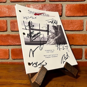 Op de afbeelding: Een gesigneerd script van "Sweeney Todd: The Demon Barber of Fleet Street" op een houten standaard. Het script bevat een zwart-wit afbeelding en meerdere handtekeningen. De achtergrond is een bakstenen muur.