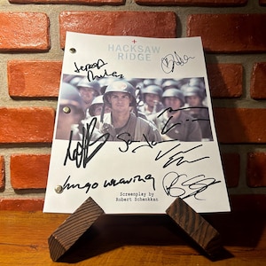 May include: A signed script from the movie "Hacksaw Ridge" featuring a photo of soldiers. The script is propped up on a wooden stand against a brick wall. The title and screenplay credits are visible.