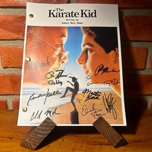 May include: A signed print of "The Karate Kid" movie poster, featuring the film's title and credits. The poster displays images of the main characters and is supported by a wooden stand. The print is signed by multiple people.