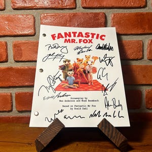 May include: A script for "Fantastic Mr. Fox" with signatures, displayed on a wooden stand. The title is in red, and the script features illustrations of fox characters. The background is a brick wall.