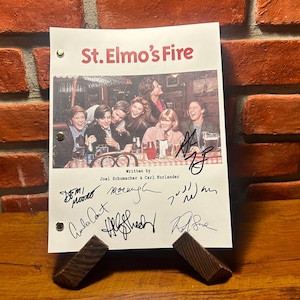 May include: A signed script from the film "St. Elmo's Fire" with a colour photo of the cast. The title is in red, and the script is supported by two wooden stands. The background is a brick wall.