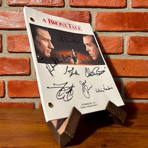 May include: A script for "A Bronx Tale" with actor signatures, displayed on a wooden stand. The script features images of Robert De Niro and Chazz Palminteri. The background is a brick wall.