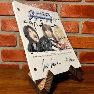 Puede incluir: Un guion blanco con el nombre de la banda "Spinal Tap" e imágenes de los miembros de la banda. El guion está firmado y se muestra en un soporte de madera. El fondo es una pared de ladrillos.