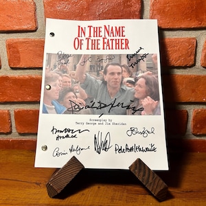 May include: A signed script from the film "In the Name of the Father" is displayed on a wooden stand. The script features the film's title in red and includes signatures. The image shows a scene from the movie.