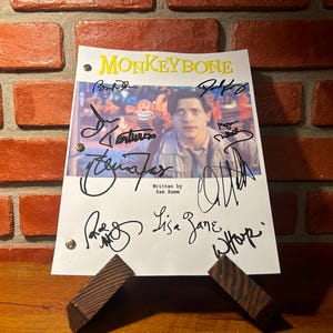 May include: A white script with the title "Monkeybone" and signatures, propped up on a wooden stand. The script features a movie still with a man and a cartoon monkey. The background is a brick wall.