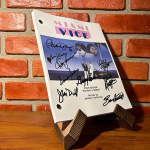 Könnte beinhalten: Ein Drehbuch für die Fernsehsendung "Miami Vice" auf einem Holzständer. Das Titelblatt des Drehbuchs zeigt das Logo der Sendung und ein Foto der Darsteller. Das Drehbuch ist von mehreren Personen signiert. Die Pilotfolge trägt den Titel "Brother's Keeper".