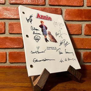 May include: A white script with the title "Annie" and signatures, supported by a wooden stand. The script features an illustration of a girl and a dog. The background is a brick wall.