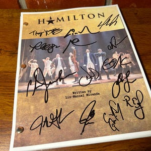 May include: A signed "Hamilton" playbill with multiple signatures in black ink. The playbill features a stage photo of the cast and the text "Written by Lin-Manuel Miranda" at the bottom. The background is a wooden surface.