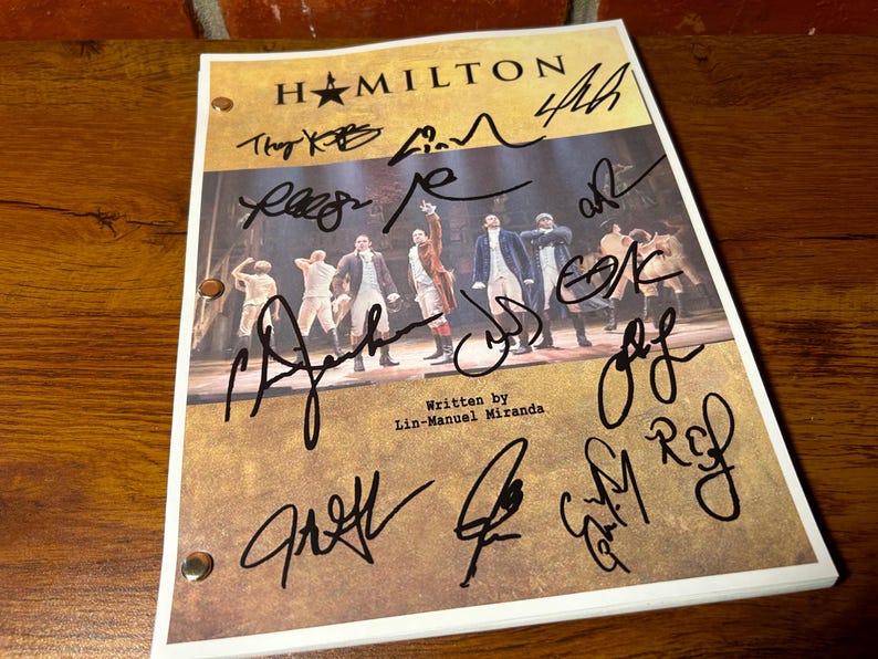 May include: A signed "Hamilton" playbill with multiple signatures in black ink. The playbill features a stage photo of the cast and the text "Written by Lin-Manuel Miranda" at the bottom. The background is a wooden surface.