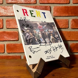 May include: A script for the musical "RENT" with the title in colourful letters. The script is signed by multiple people and is displayed on a wooden stand. The background is a brick wall.