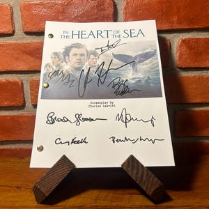 Könnte beinhalten: Ein signiertes Drehbuch für "Im Herzen der See" mit Chris Hemsworth. Das Filmposter zeigt eine Walflosse und Schauspieler. Das Skript wird von einem Holzständer vor einer Backsteinmauer gehalten.